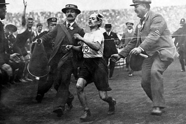 Dorando Pietri sendo ajudado na linha de chegada por fiscais da prova para ficar em primeiro lugar na maratona de 1908.